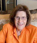 An older woman in an orange shirt sitting on a couch.